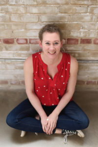 Alyssa Alexander is a white woman with short blond hair. She is sitting cross-legged on the floor with a brick wall behind her.