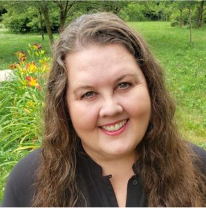 Susan Gee Heino is a white woman with long brown hair with gentle waves. She's sitting in front of some orange daylilies.