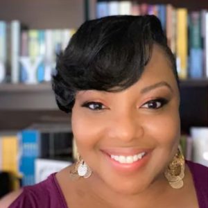 Otescia Johnson is a black woman with short, black hair. She's wearing a magenta blouse, and is standing in front of a bookcase.