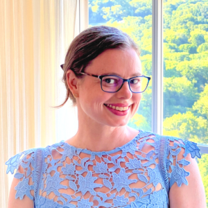 Libby Waterford is a white woman. Her brown hair is tied back behind her head, and she is wearing glasses and a lacy blue top. 