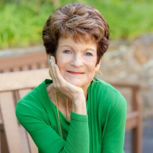 DiAnn Mills is a white woman with short-cropped reddish brown hair. She's wearing a green long-sleeved top and sitting on a bench.