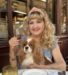 Teri Wilson is a white woman with long, gently curling blond hair. She's holding a beverage. A cute brown and white dog with long brown ears that that dangle down like hair sits in her lap.