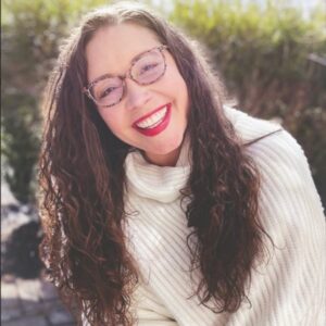Jen Devon is a white woman with long brown hair. She wears glasses as wide as her face, and is wearing an off-white sweater.